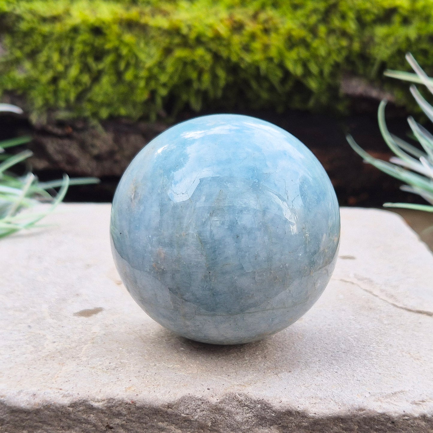 This Aquamarine Crystal Sphere features a soft, pale aqua blue tone with natural inclusions that add to its individual character. A subtle opalescent effect appears when turned in the light. You'll notice dark grey-black inclusions (possibly biotite) and soft white areas (possibly feldspar).