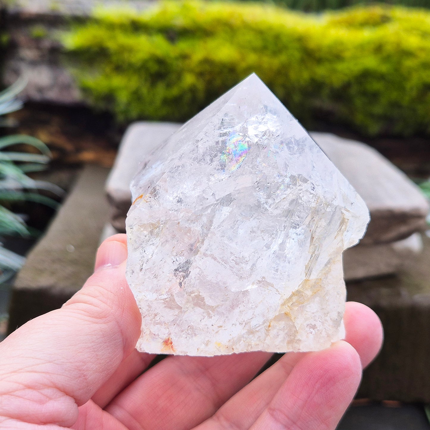 Quartz Crystal Standing Point from Brazil. This piece features a polished tip with a visible rainbow inclusion, contrasted by natural sides. The flat base allows it to stand freely, making it an ideal display crystal. It has lovely clarity, and the way it catches the light adds a beautiful reflective quality.