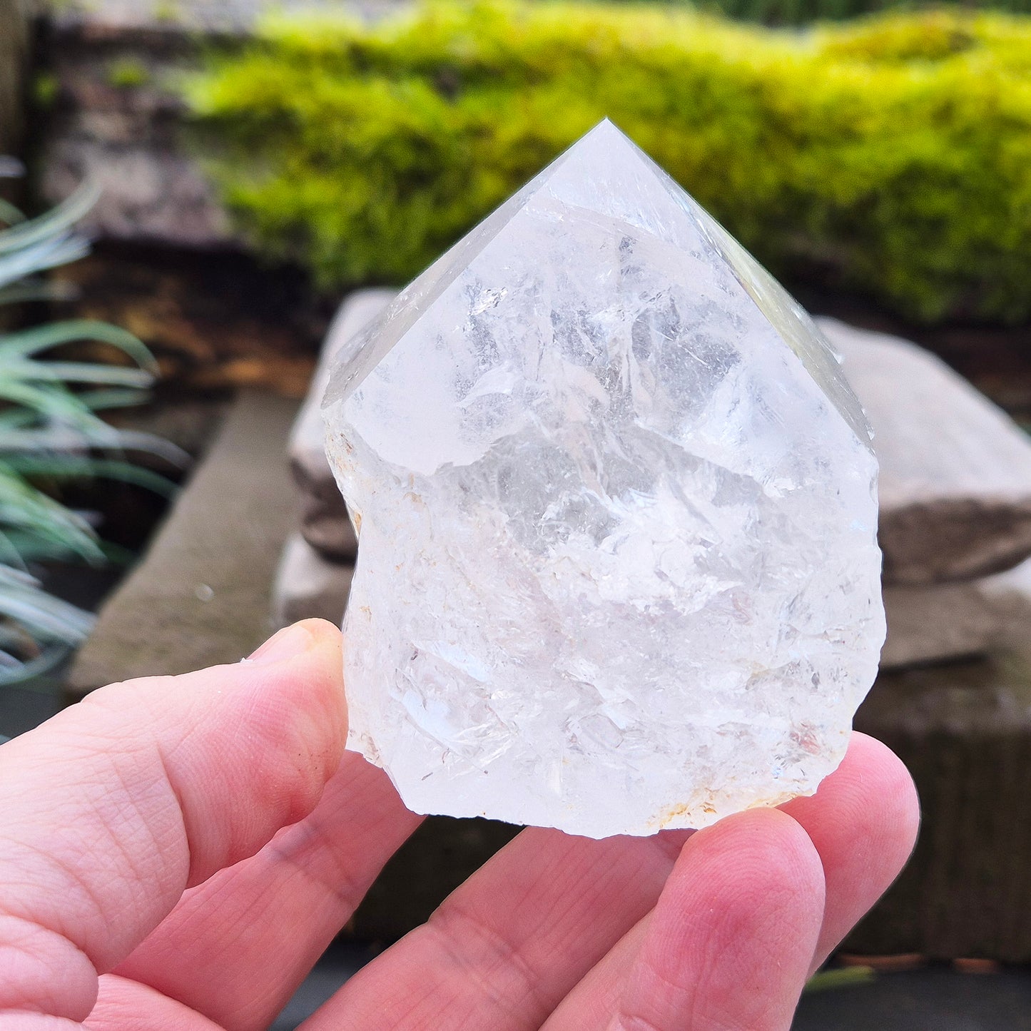 Quartz Crystal Standing Point from Brazil. This piece features a polished tip with a visible rainbow inclusion, contrasted by natural sides. The flat base allows it to stand freely, making it an ideal display crystal. It has lovely clarity, and the way it catches the light adds a beautiful reflective quality.