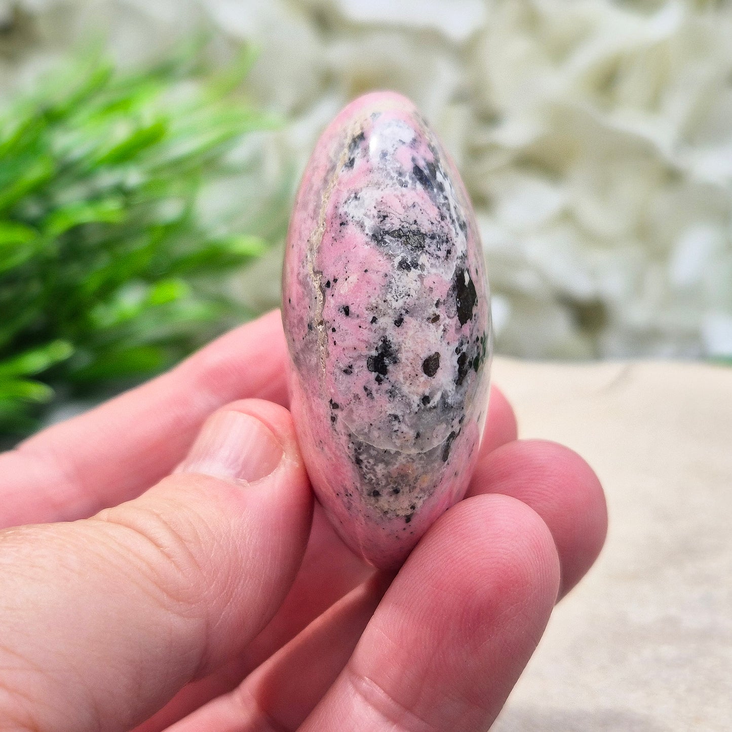 Pink Rhodonite Crystal Palm Stone – also known as Bubblegum Pink Rhodonite from Peru. This beautiful Rhodonite crystal, sometimes nicknamed Bubblegum Pink Rhodonite, comes from Peru and features a soft pink colour with black patches caused by the natural oxidation of manganese.