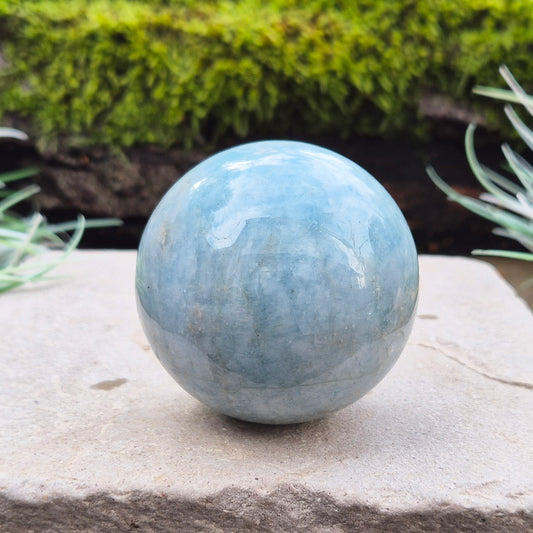 This Aquamarine Crystal Sphere features a soft, pale aqua blue tone with natural inclusions that add to its individual character. A subtle opalescent effect appears when turned in the light. You'll notice dark grey-black inclusions (possibly biotite) and soft white areas (possibly feldspar).