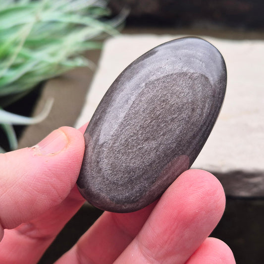 Silver Sheen Obsidian Palm Stone is a volcanic glass with a striking silvery or metallic sheen, created by tiny gas bubbles trapped during rapid lava cooling.