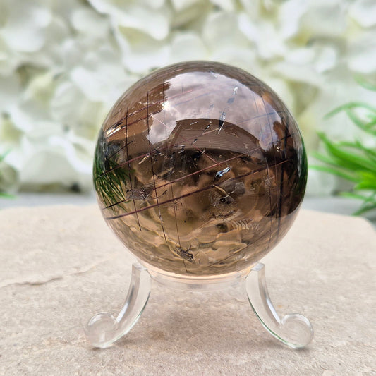Smoky Quartz Sphere with Copper Red Rutile and rainbow flashes — grounding, protecting, and boosting creativity and clarity. Includes acrylic stand.