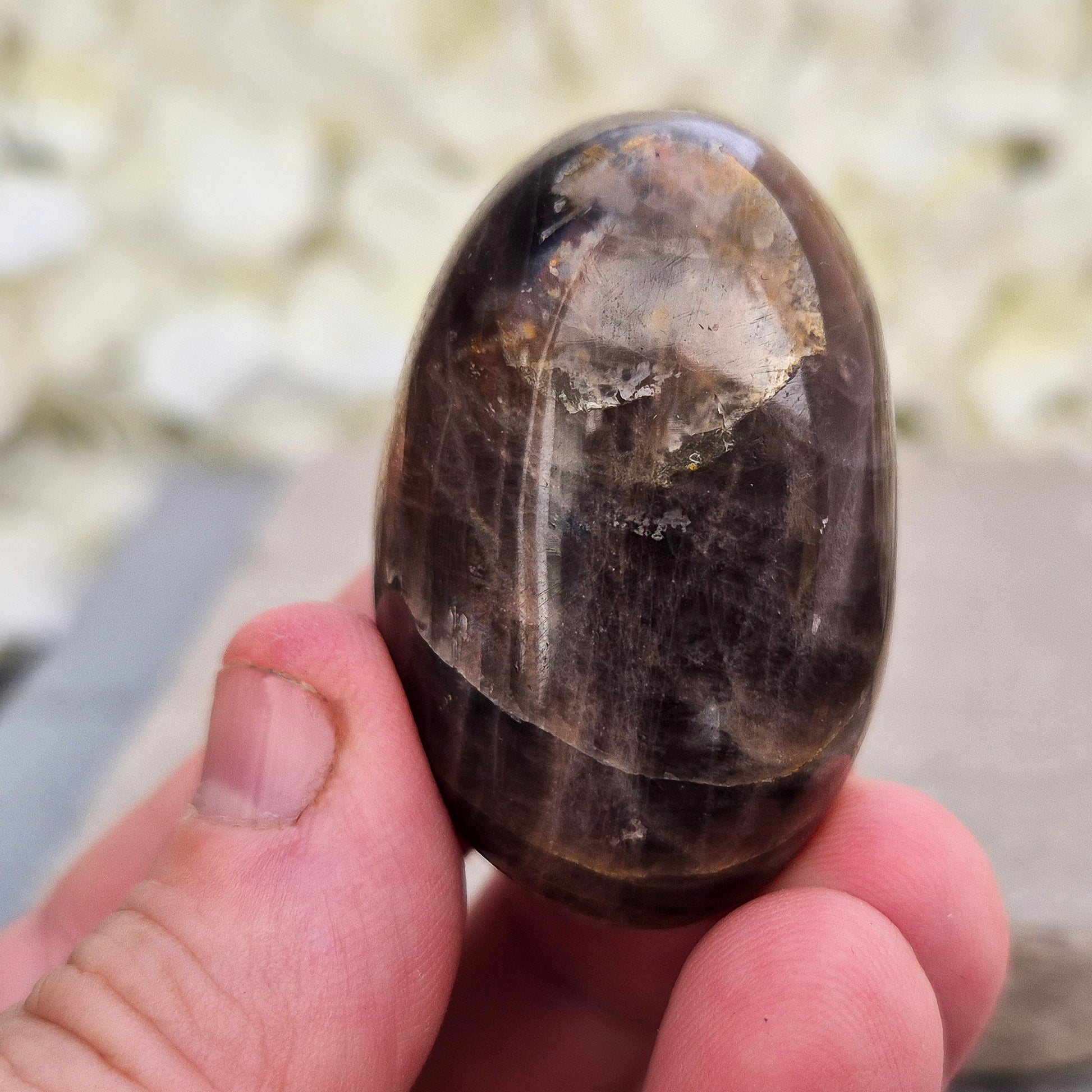 Black Moonstone Crystal Palm Stone. This polished Black Moonstone palm stone from Madagascar features deep shades of gray, black, and brown with subtle flashes of silver, peach, or white. Its smooth surface fits comfortably in your hand, offering a grounding and calming presence.