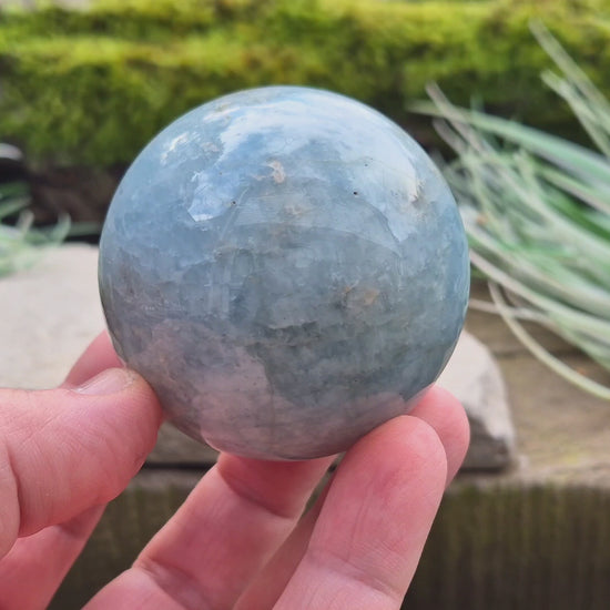 This Aquamarine Crystal Sphere features a soft, pale aqua blue tone with natural inclusions that add to its individual character. A subtle opalescent effect appears when turned in the light. You'll notice dark grey-black inclusions (possibly biotite) and soft white areas (possibly feldspar).