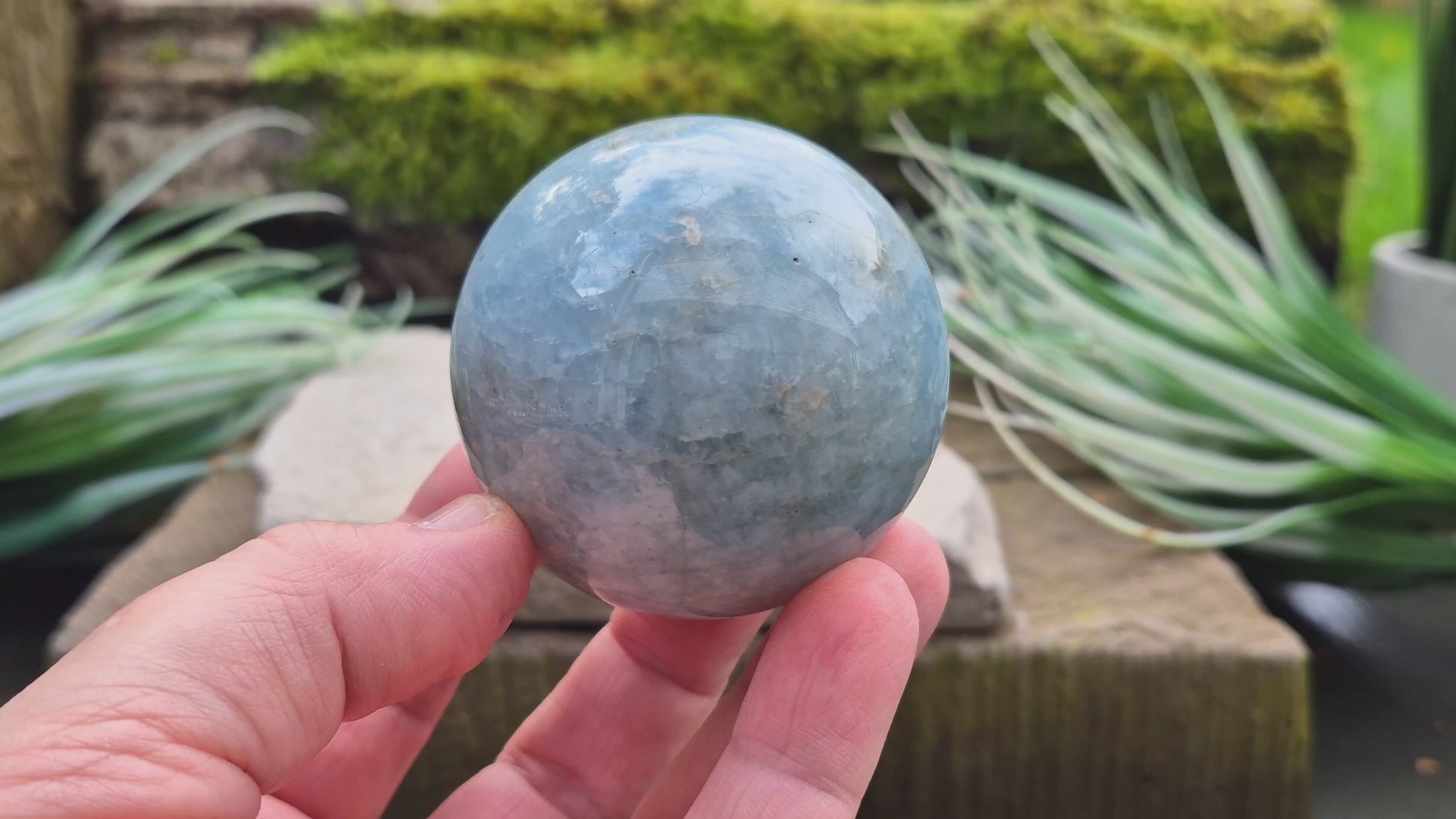 This Aquamarine Crystal Sphere features a soft, pale aqua blue tone with natural inclusions that add to its individual character. A subtle opalescent effect appears when turned in the light. You'll notice dark grey-black inclusions (possibly biotite) and soft white areas (possibly feldspar).