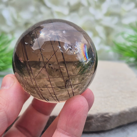 Smoky Quartz Sphere with Copper Red Rutile and rainbow flashes — grounding, protecting, and boosting creativity and clarity. Includes acrylic stand.
